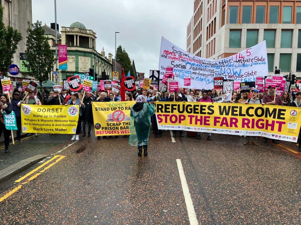 a crowd shot of the protest against the Tory party conference, people stand behind a big yellow banner that reads refugees welcome here